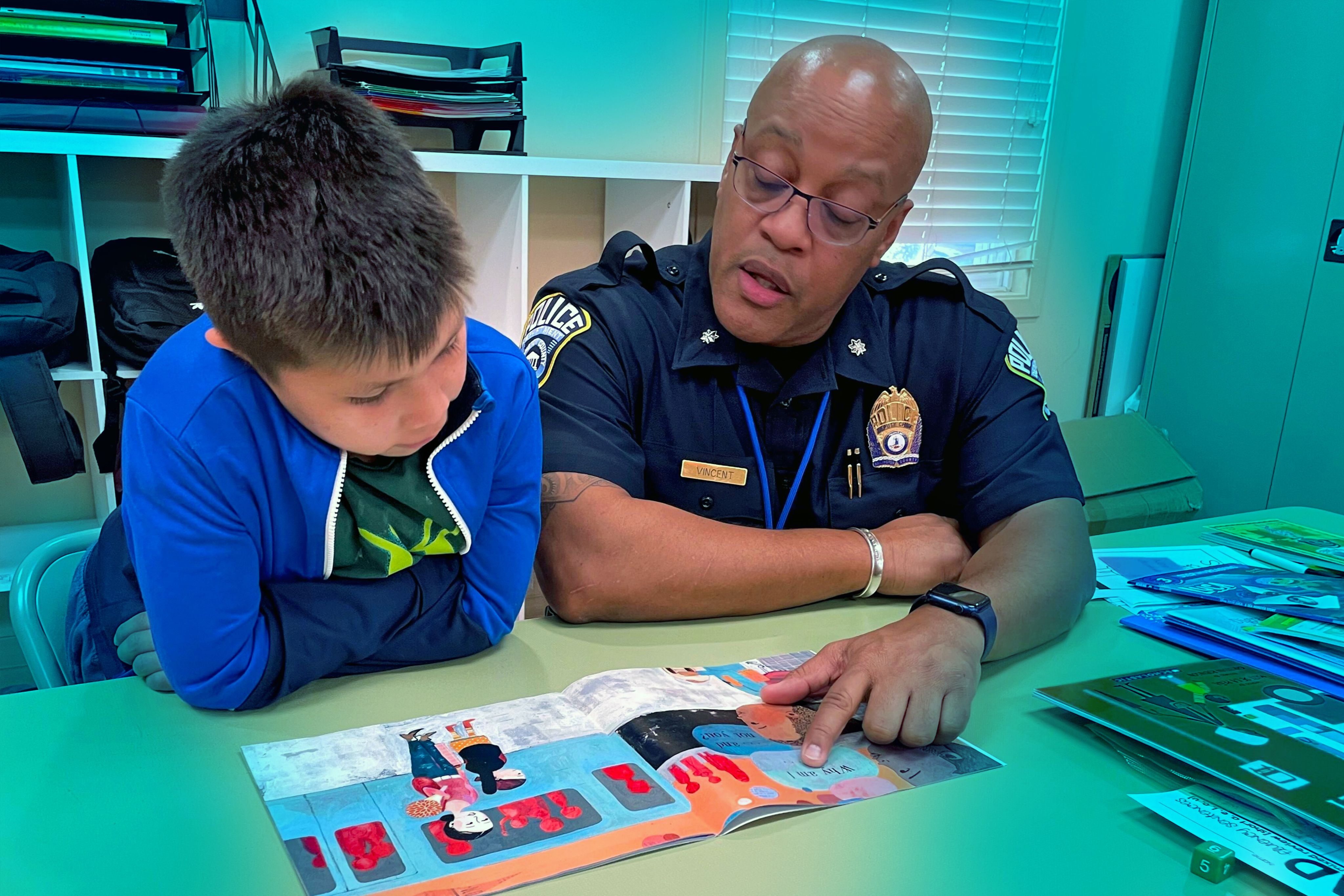 An Arlington County police officer points at a book and reads with a young student.