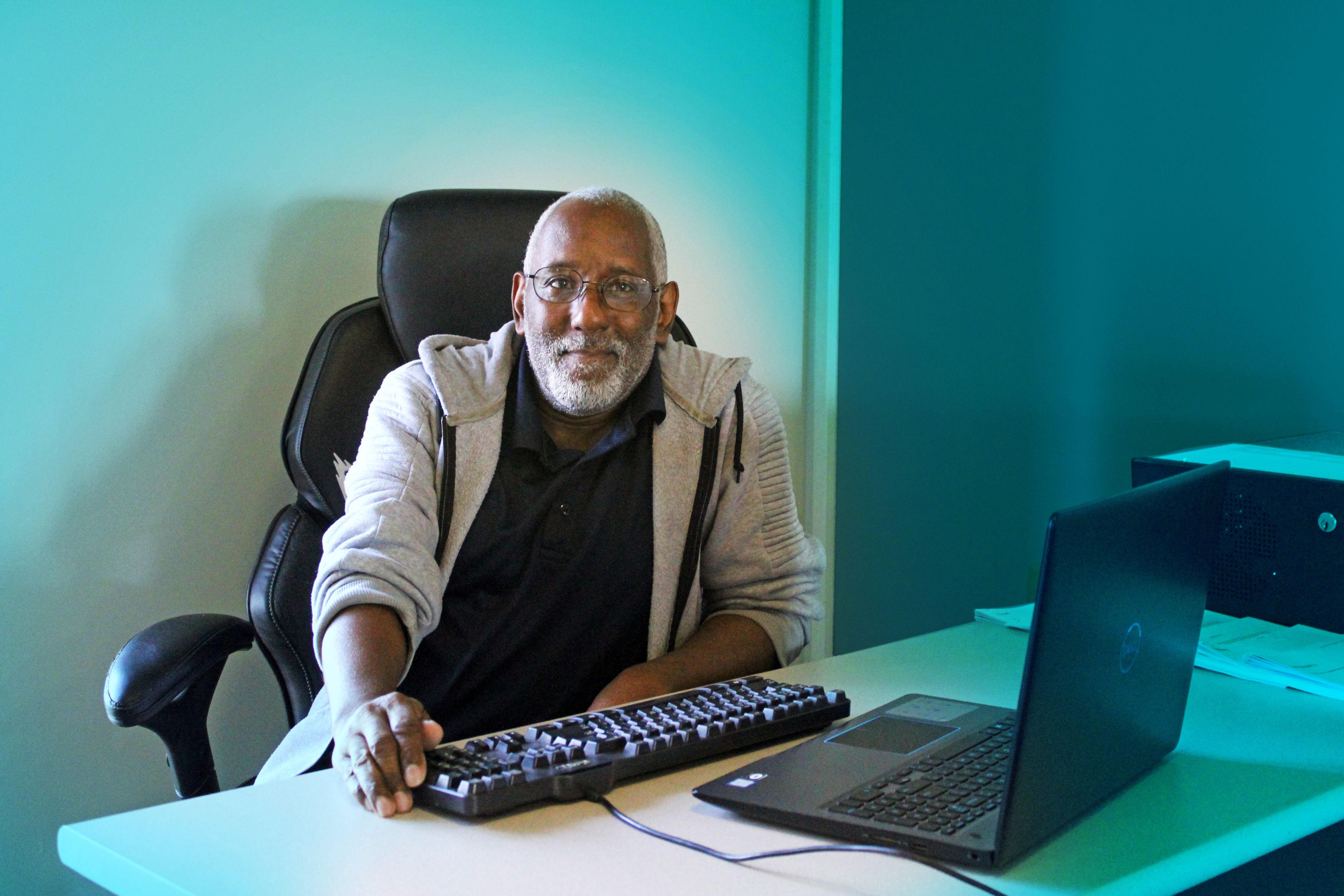 An older man has a soft smile and is sitting at a desk with a keyboard and a computer.
